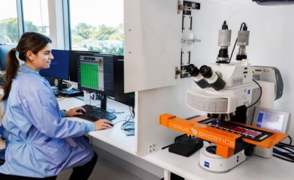 A scientist in SNP's Brisbane laboratory beside one of the digital pathology scanners. A female scientist in a blue lab coat sits at a computer beside a digital microscope scanning system.
